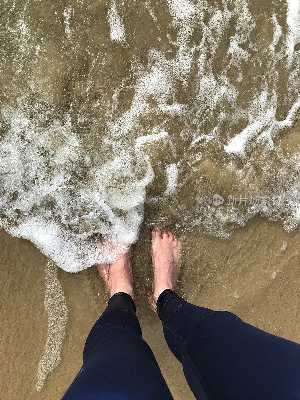 两只白色的脚在沙滩上，一个英国男人正在弄湿深蓝色的男士紧身裤/运动慢跑裤，在海浪中划桨/站立的脚印冲刷着金色的沙滩，光着脚在海边的海浪中沉入潮湿的沙滩